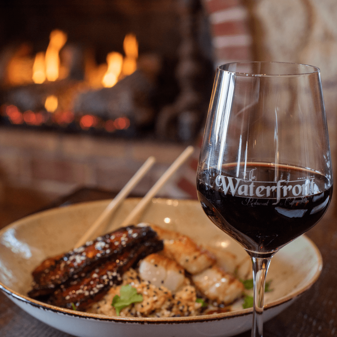 A glass of red wine beside a seafood dish with a fireplace in the background.